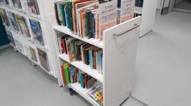 Valkoinen kirjakärry kirjaston kirjahyllyn päädyssä täynnä kirjoja ja kyltti jossa lukee kirjojen kierrätyskärry.