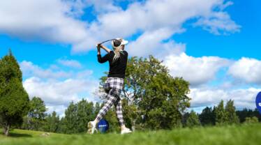 Aurinkoisella säällä golffaava nainen jolla on musta-valko ruudulliset housut ja musta paita sekä vaaleat pitkät hiukset.
