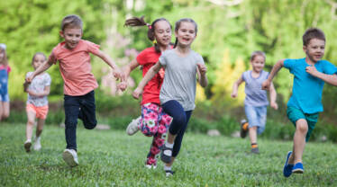 Iloiset lapset juoksevat nurmikentällä taustalla lehtipuita kesällä.