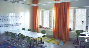 Valkoisella tussitaululla kirjaintauluja, edessä pöytä jonka ääressä 8 tuolia, taustalla ikkunoita ja oransseja verhoa.