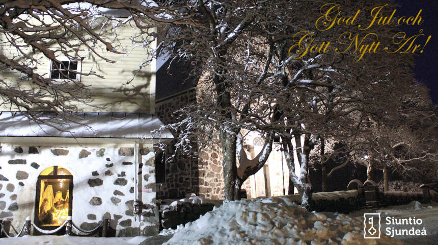 Kyrkan och klockstapeln med upplyst julkrubba i fönstret i mörkret med mycket snö på marken och i träden.