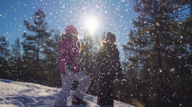 Kaksi lasta lumisessa rinteessä, taustalla aurinko, edessä pyryää lumihiutaleita.