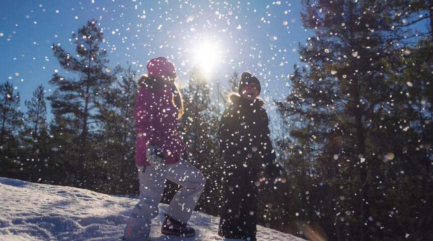 Kaksi lasta lumisessa rinteessä, taustalla aurinko, edessä pyryää lumihiutaleita.